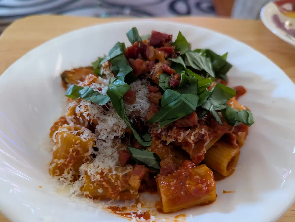A close-up photo of a white bowl filled with rigatoni pasta in a red tomato sauce, topped with crumbled bacon, fresh basil…