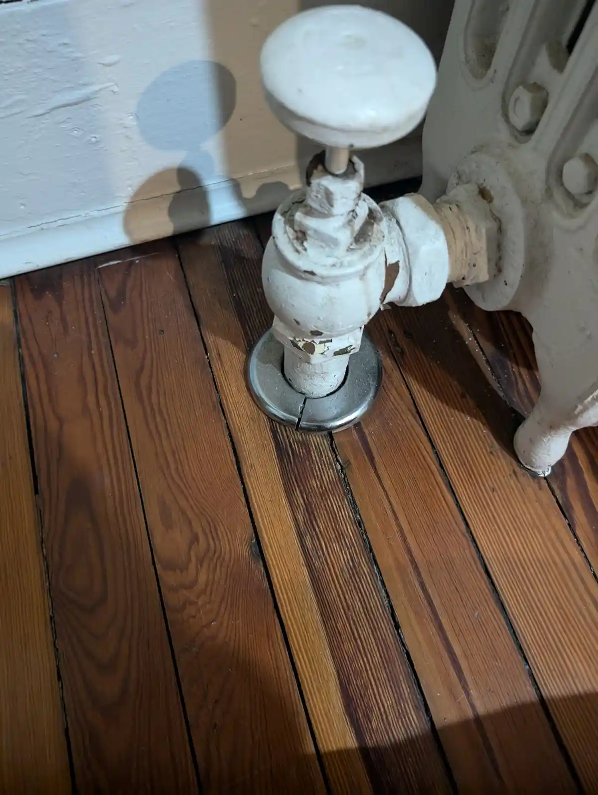 A close-up photo shows a weathered, white-painted radiator valve with peeling paint, mounted on a wooden floor next to a…