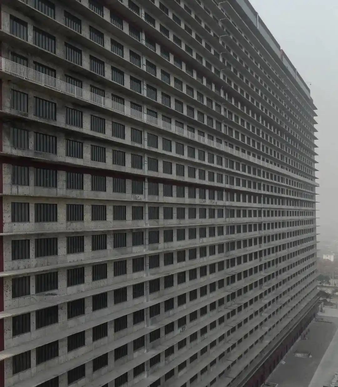 A tall, unfinished concrete building with repetitive rows of window openings and balconies, viewed from an angle against a…