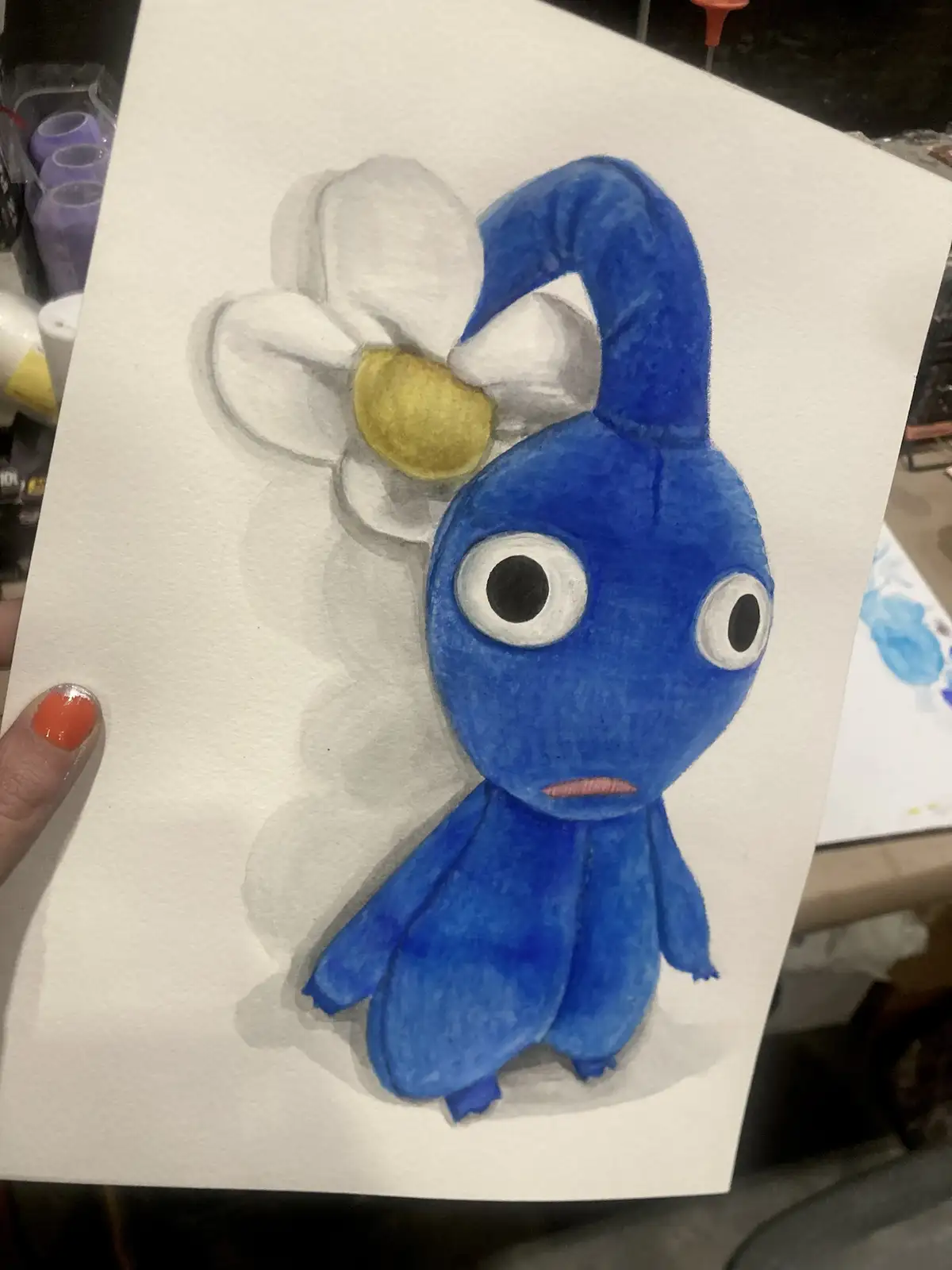 A hand holds up a watercolor drawing of a blue, round, cartoonish creature with large eyes and a small mouth, resembling a…