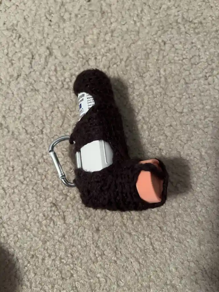 A close-up photo shows an asthma inhaler wrapped in a dark brown knitted cover, with a metal carabiner clipped to its side…