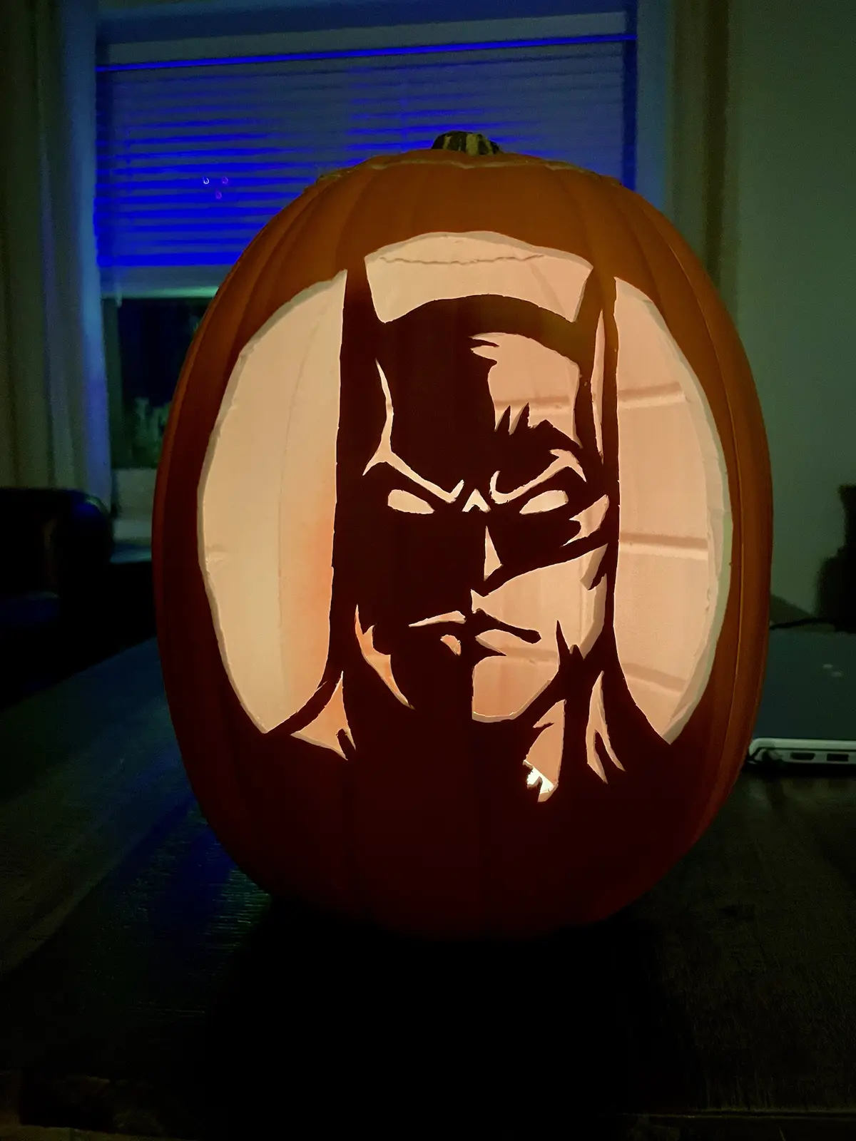 A carved Halloween pumpkin with an illuminated silhouette of Batman's face, featuring his iconic cowl and pointed ears, set…