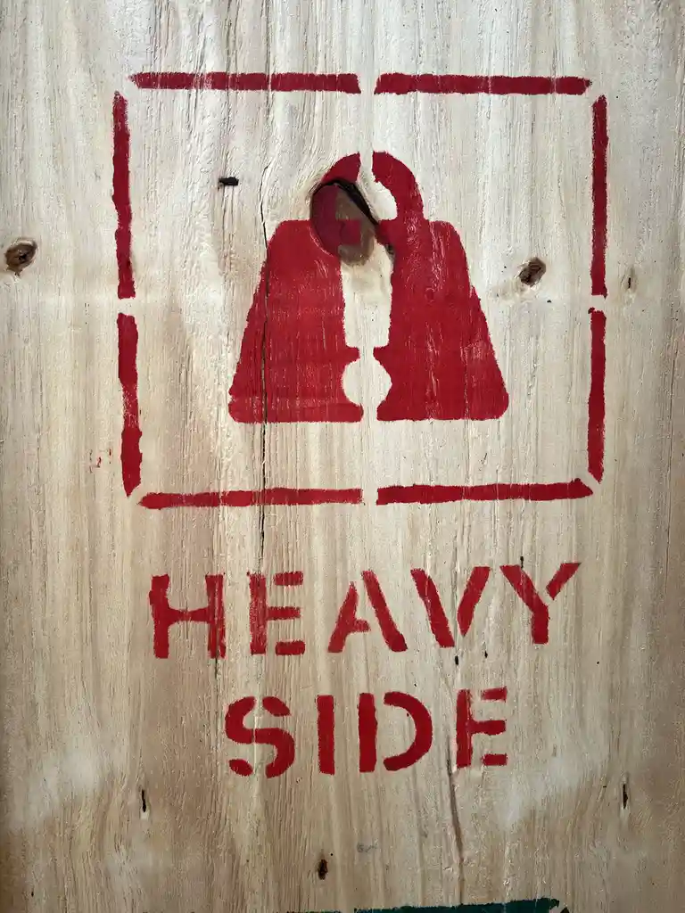 A close-up of a red-painted wooden sign showing two weight symbols inside a square, with the words 'HEAVY SIDE' below. The…
