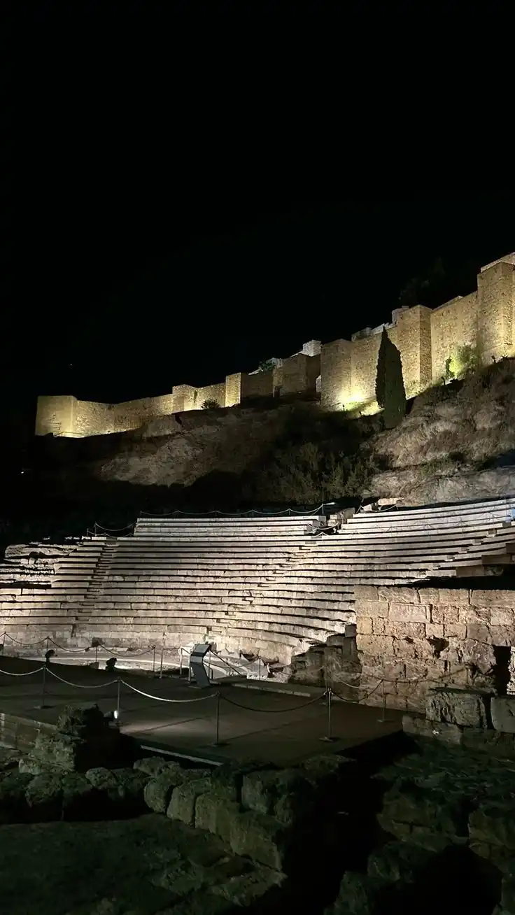A nighttime photo of an ancient stone amphitheater with tiered seating, illuminated by spotlights against a dark sky. The…