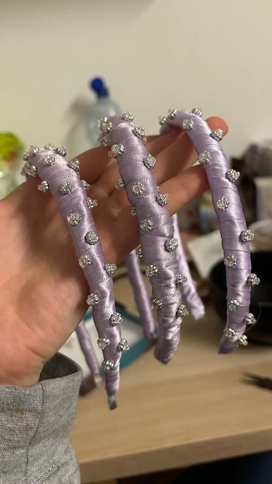 A hand holds three handmade, lavender-colored satin headbands, each decorated with numerous small, sparkling rhinestones…