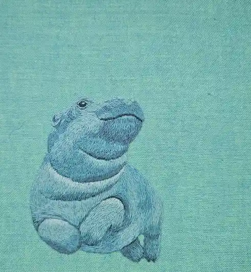 An embroidered illustration of a hippopotamus stitched in shades of blue onto a light blue fabric background. The hippo is…