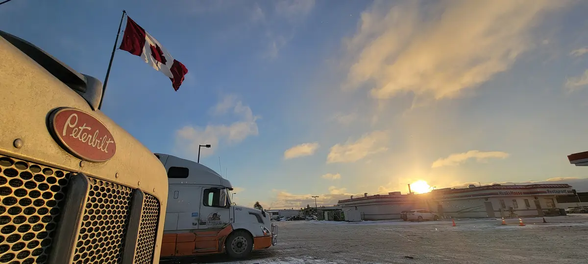 A close-up of a Peterbilt semi-truck's front grille with a Canadian flag waving on top, parked in a snowy lot at sunset. In…