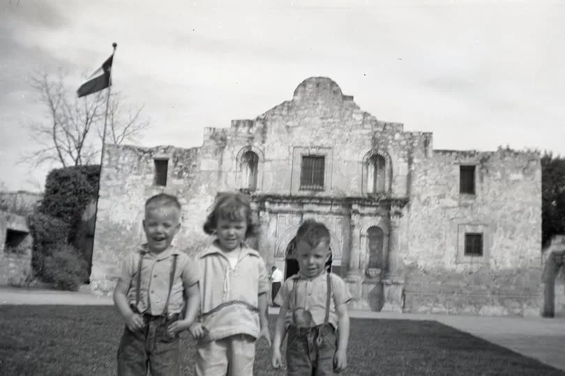 A vintage black-and-white photo shows three young children standing in front of the historic Alamo in San Antonio, Texas…