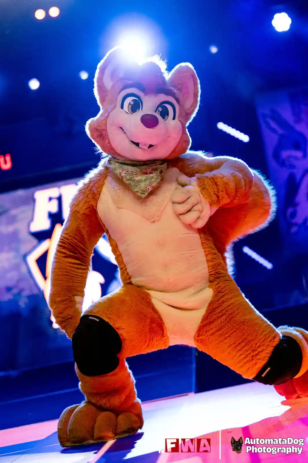 A person in a detailed orange and cream fox mascot costume poses confidently on a stage under bright blue and white lights…