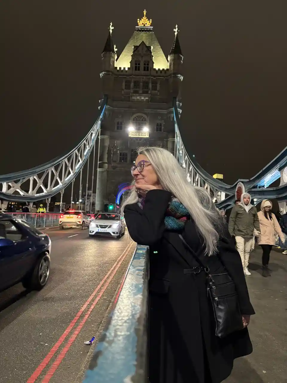 A woman with long silver hair and glasses stands on the pedestrian walkway of Tower Bridge at night, looking thoughtfully…