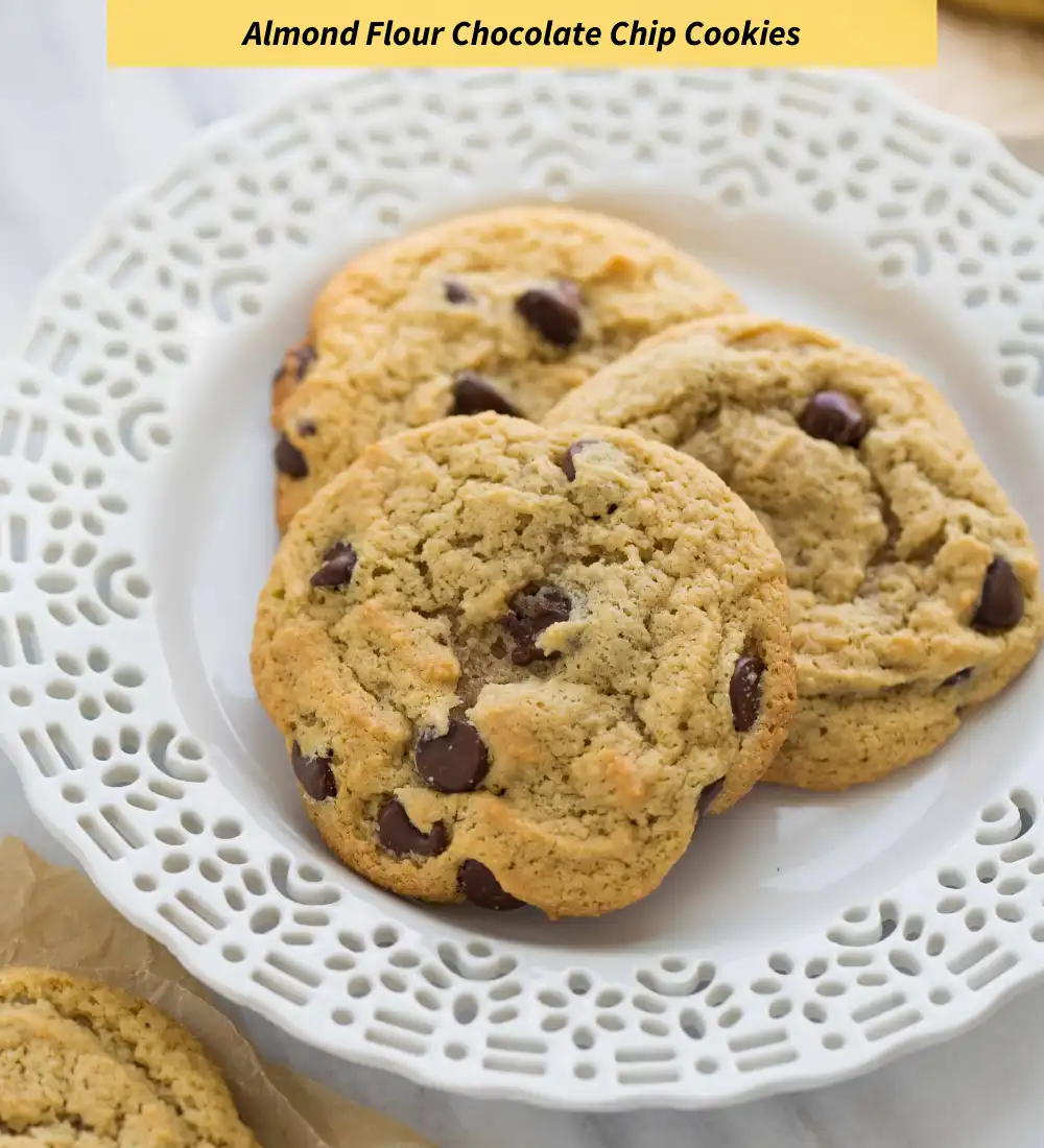 Three golden-brown chocolate chip cookies sit on a white, ornate lace-patterned plate. The cookies have visible chocolate…