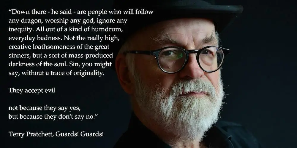 A close-up portrait of Terry Pratchett, an older man with a white beard, glasses, and a black hat, against a dark…
