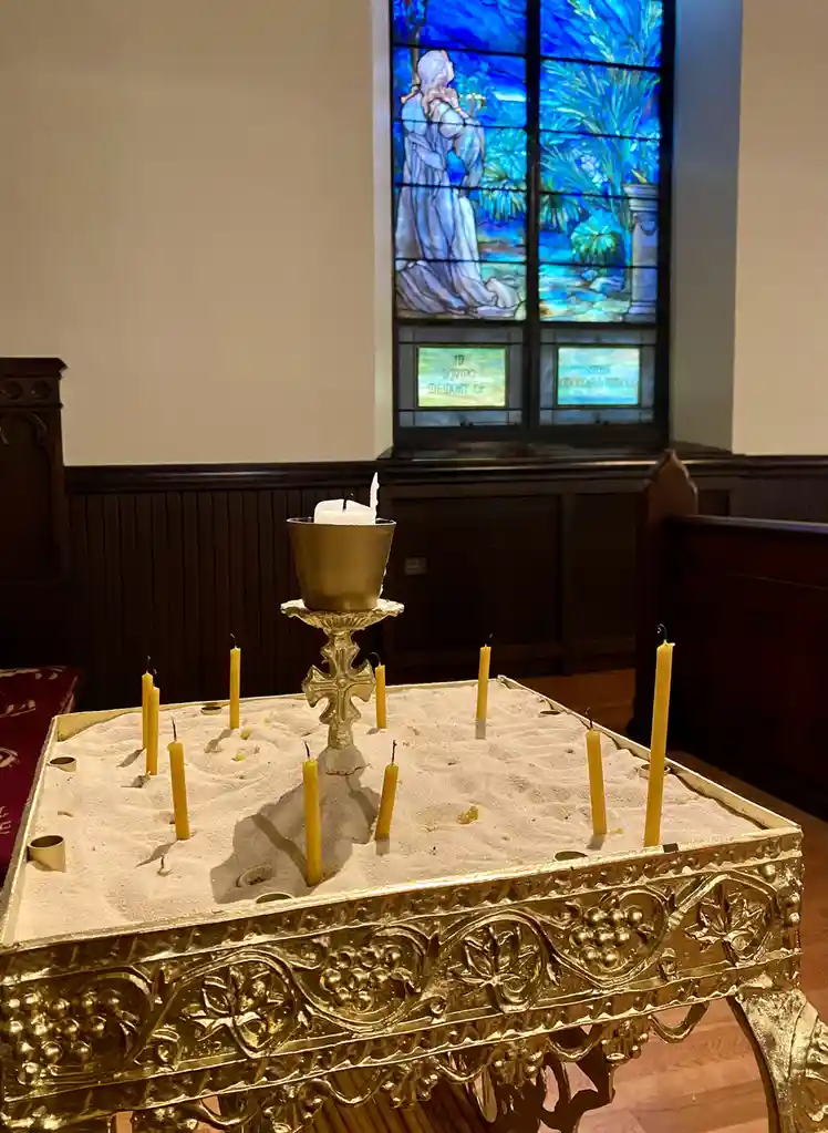 A golden ornate table covered in sand holds numerous yellow candles, some lit, with a central candle in a brass holder…