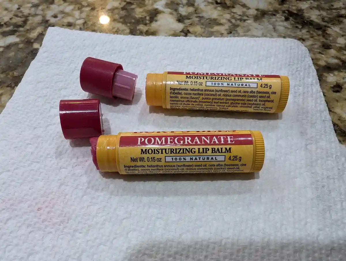 Two tubes of Burt's Bees Pomegranate Moisturizing Lip Balm lie on a white paper towel, one with its cap off and the balm…