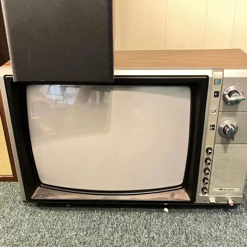 A vintage Midland CRT television with a wooden top and silver casing sits on a green carpet, its screen blank and…