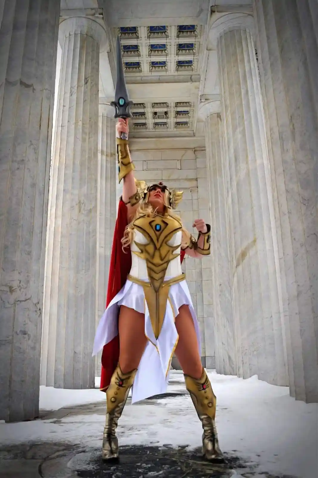 A person in a detailed, gold-and-white fantasy warrior costume with a red cape and ornate boots stands in a snowy, columned…