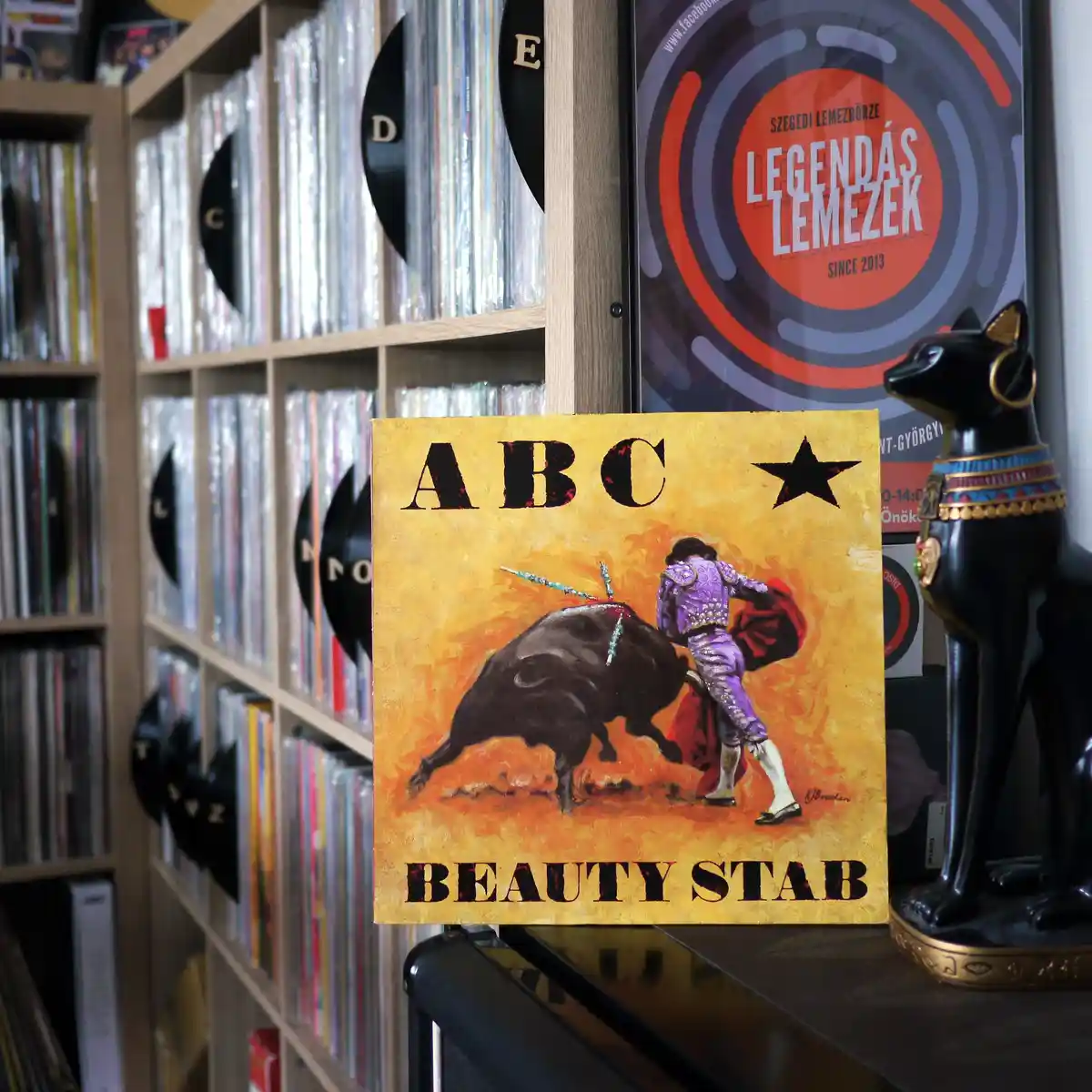 A painting of a matador in purple traje de luces facing a charging bull, with the text 'ABC' at the top and 'BEAUTY STAB' at…