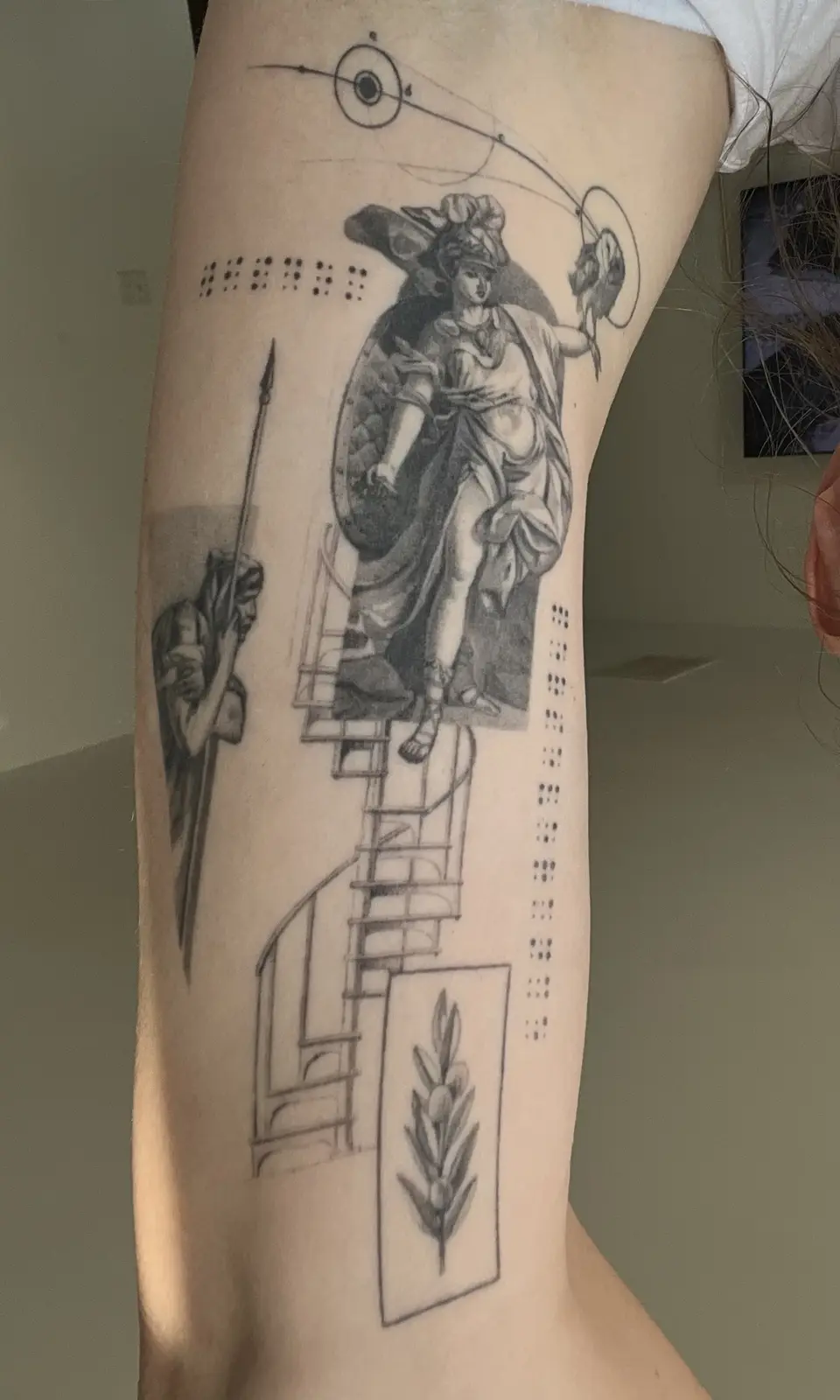 A black-and-white tattoo on a person's thigh features a classical warrior woman holding a shield and spear, standing atop a…