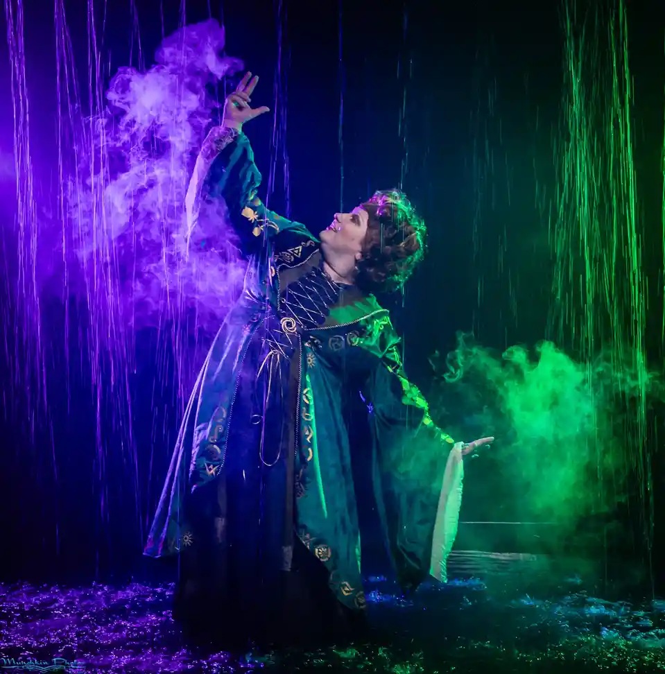 A person in a dark, ornate robe with gold trim stands in a theatrical setting, arms outstretched amidst purple and green…