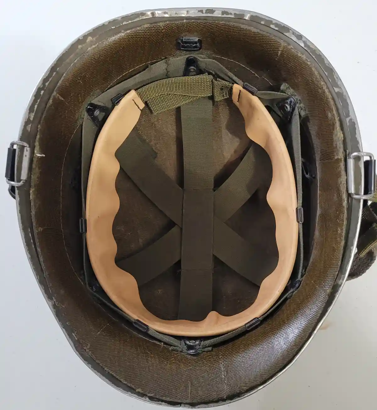 A top-down view of the inside of a worn military-style helmet, showing its olive drab fabric lining, tan padded inner rim…