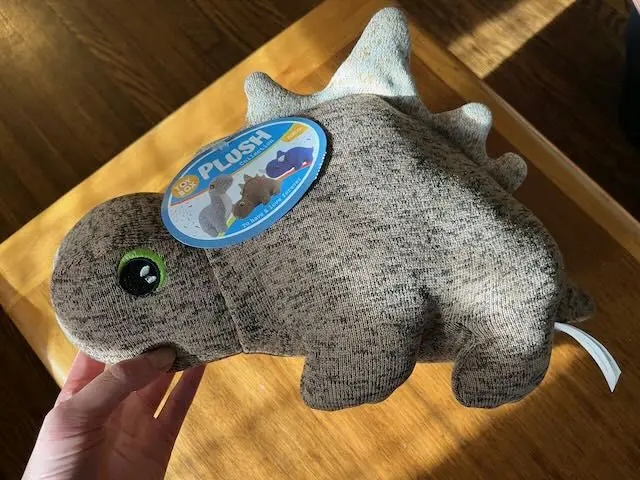 A hand holds a grey, textured plush dinosaur toy with a large green eye and a blue circular tag that reads 'Plush' and shows…