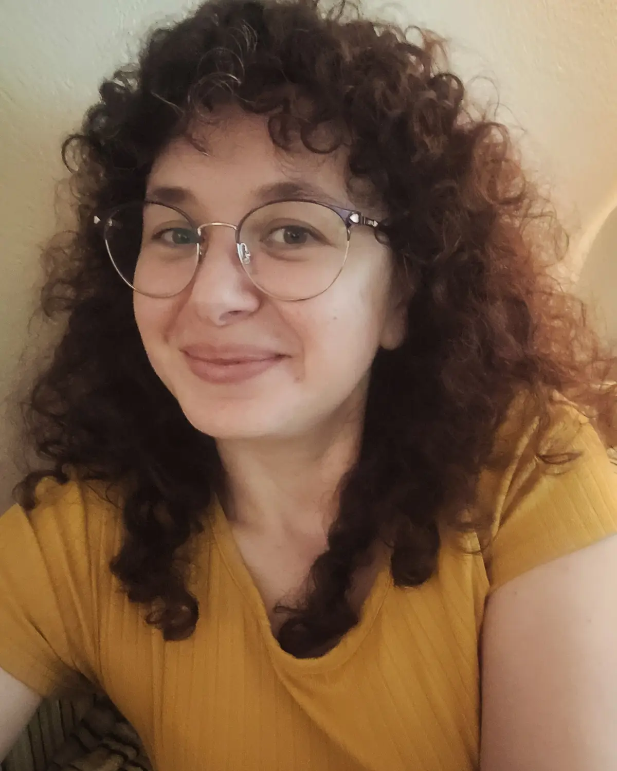 A selfie of a person with curly brown hair and glasses, wearing a yellow shirt, smiling slightly at the camera. The overlaid…