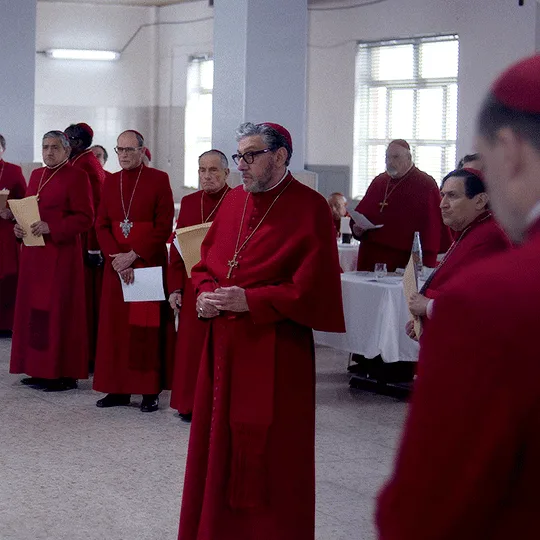 A sequence of images shows a man in a red clerical robe standing among other similarly dressed men in a room with white…
