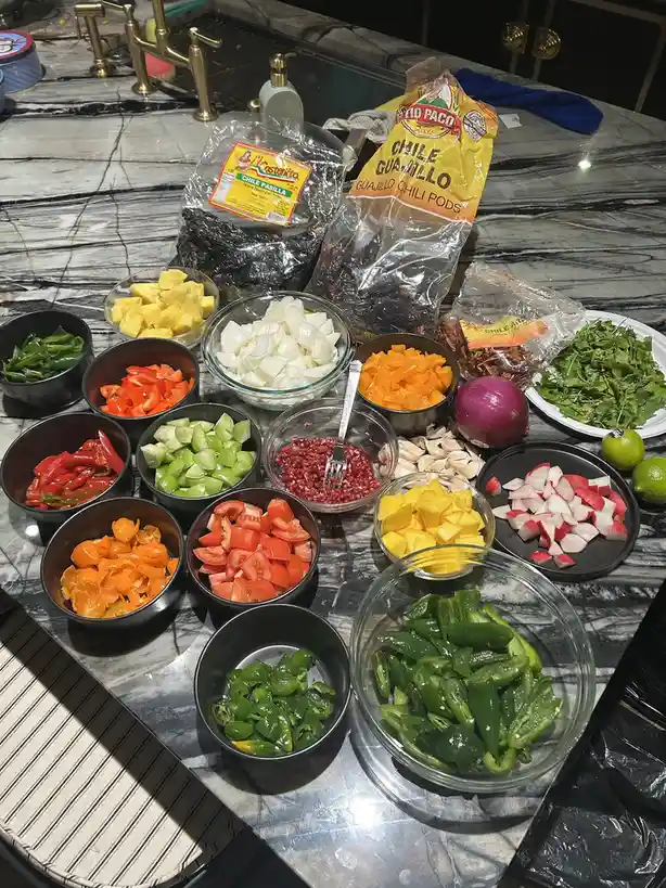 A kitchen counter is covered with numerous bowls of chopped vegetables and dried chiles, including tomatoes, peppers…