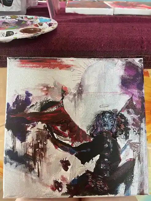 A close-up photo shows a hand holding a small canvas painting depicting a dark, abstract figure with blue skin, messy hair…