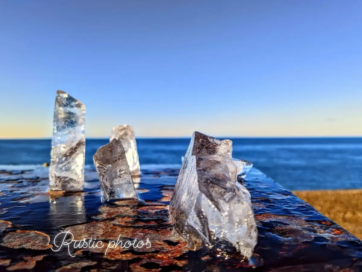 Several jagged, transparent ice formations stand upright on a rusted, weathered metal surface near a body of water, with a…