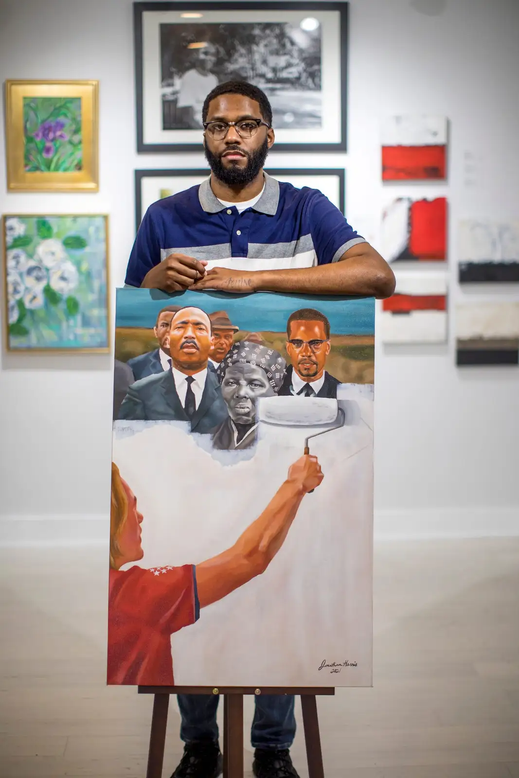 A man stands in an art gallery holding a large painting on an easel. The painting depicts historical Black figures like…