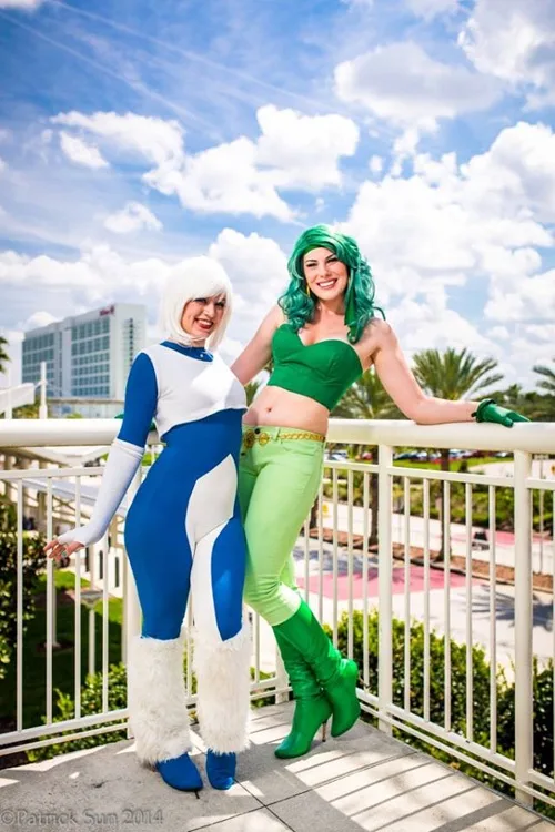 Two women in cosplay pose on a balcony under a bright blue sky with clouds. The woman on the left wears a blue and white…