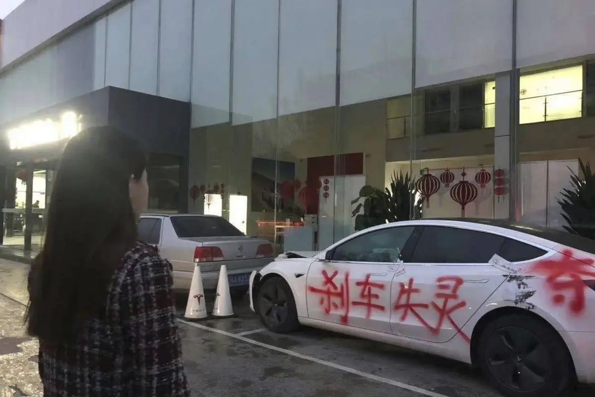 A woman stands in front of a white Tesla Model 3 that has been vandalized with red spray paint reading '刹车失灵' (brakes…