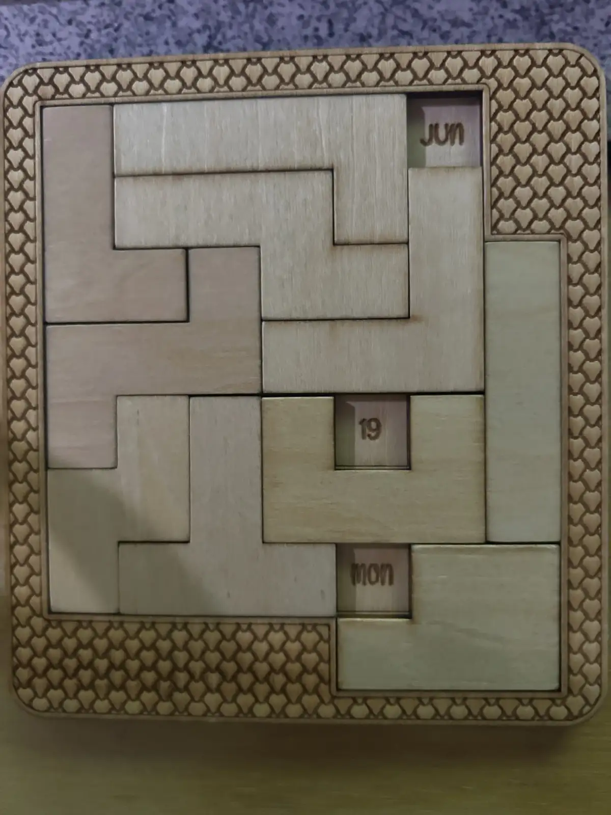 A wooden puzzle calendar with interlocking pieces forming a grid, showing 'jun' (June), '19', and 'mon' (Monday) engraved on…