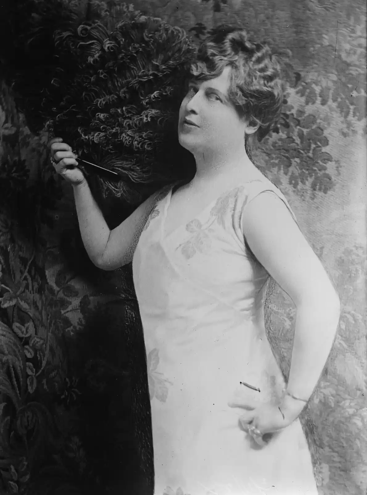 A vintage black-and-white portrait of a woman in a light-colored dress holding a large, dark feather fan. She is looking…