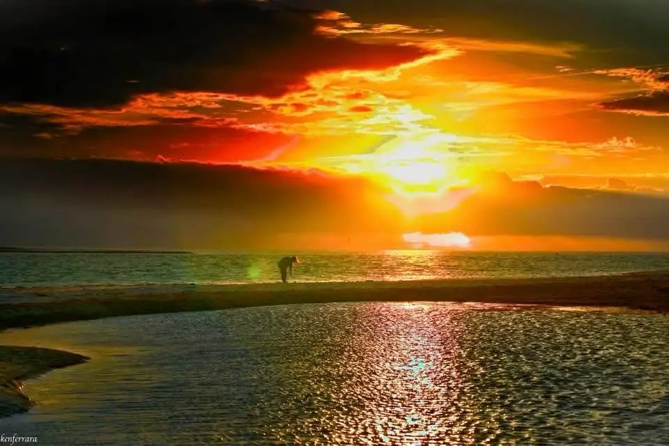 A person is silhouetted against a dramatic, fiery sunset over the ocean, bending over near the water's edge. The sky is…