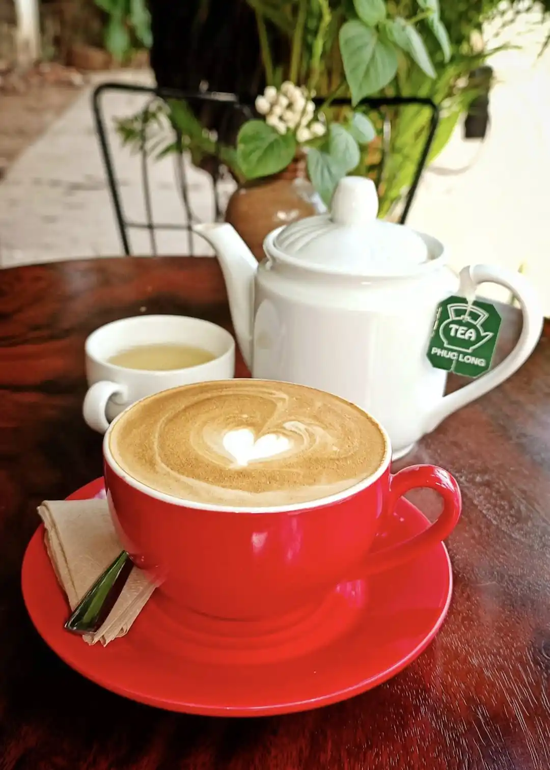 A red cup of coffee with heart-shaped latte art sits on a saucer next to a white teapot with a 'TEA PHUC LONG' tag and a…