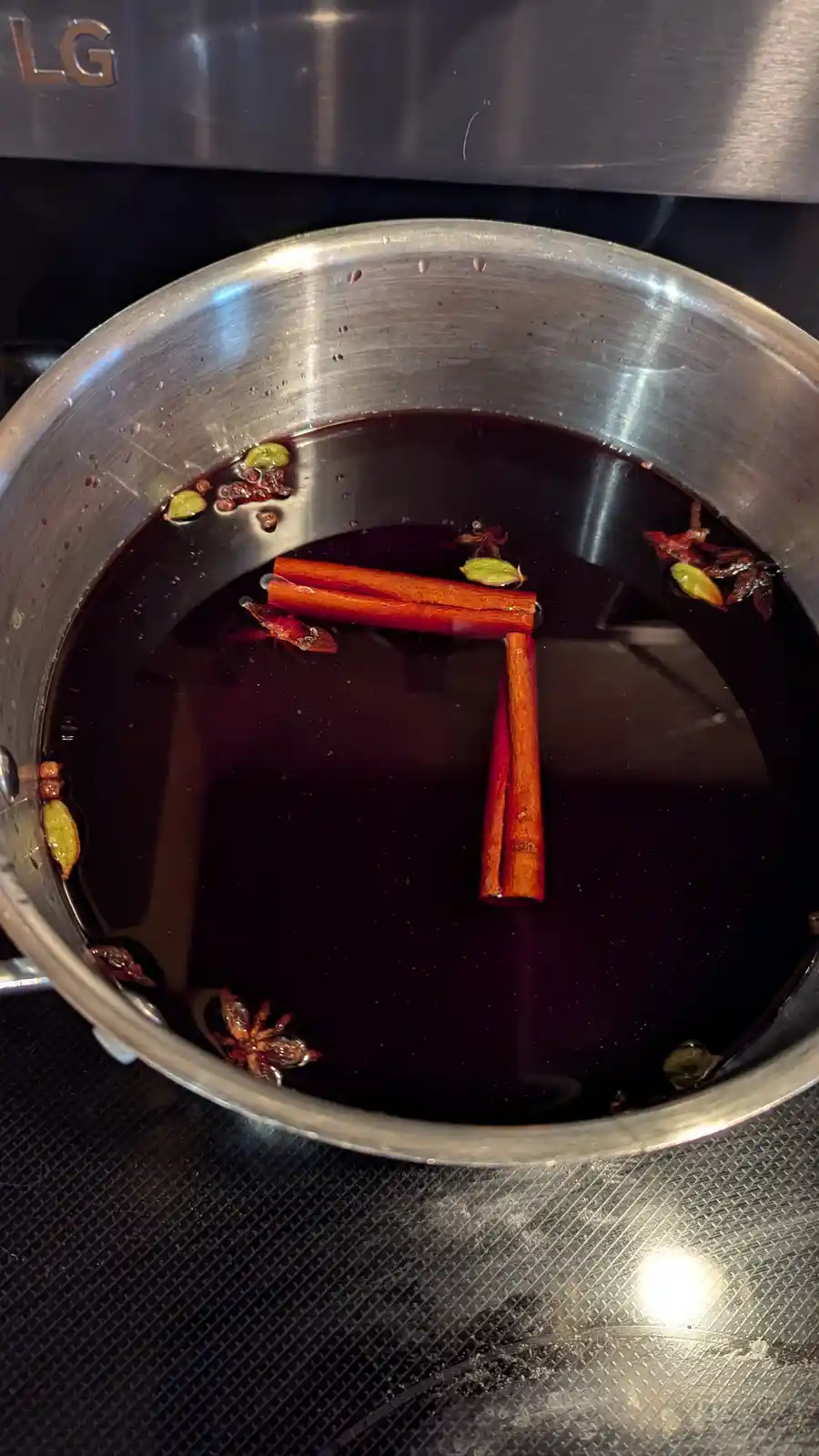A stainless steel saucepan on a stovetop contains dark liquid with floating spices including cinnamon sticks, star anise…