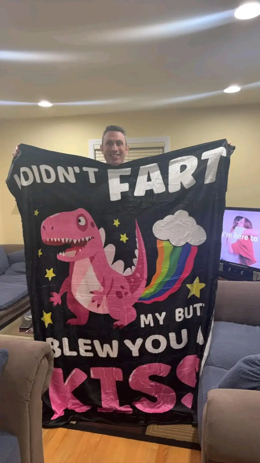 A man is smiling and holding up a black blanket featuring a cartoon pink dinosaur with a rainbow fart cloud. The blanket has…