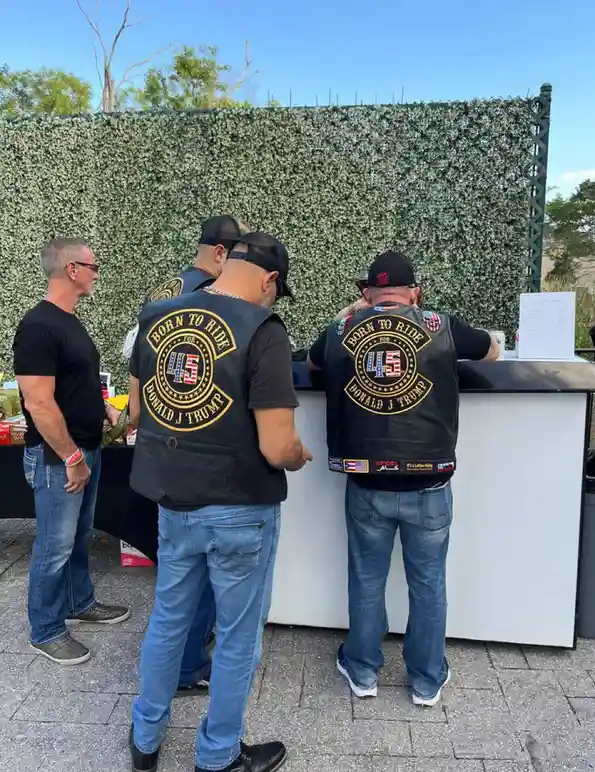 Three men stand at an outdoor counter, two wearing black vests with a circular patch that reads 'BORN TO RIDE FOR 45 DONALD…