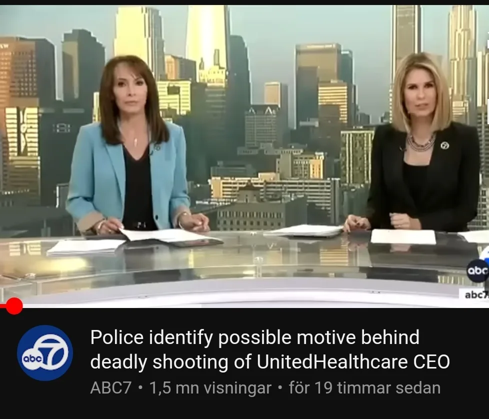 Two female news anchors sit at a desk in front of a city skyline backdrop, looking serious. A news ticker at the bottom…