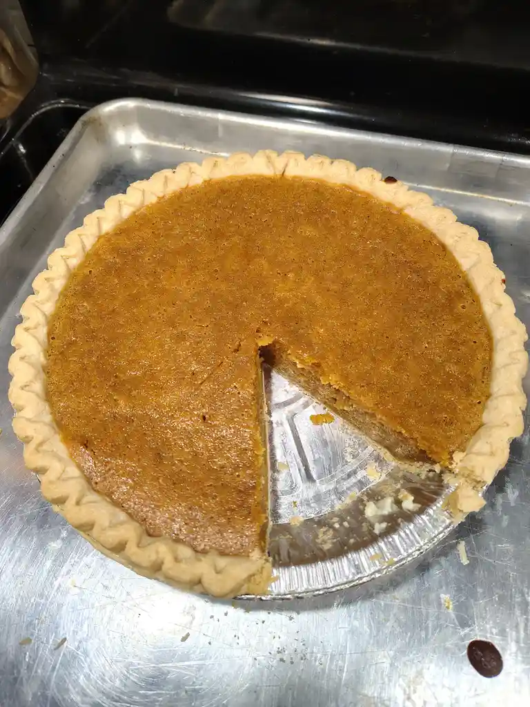 A close-up photo shows a pumpkin pie in a foil pie tin, resting on a metal baking sheet. A single slice has been removed…