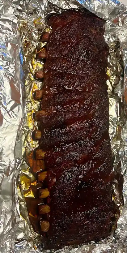 A close-up photo shows a long rack of cooked ribs covered in a dark, glossy sauce, resting on crumpled aluminum foil. The…