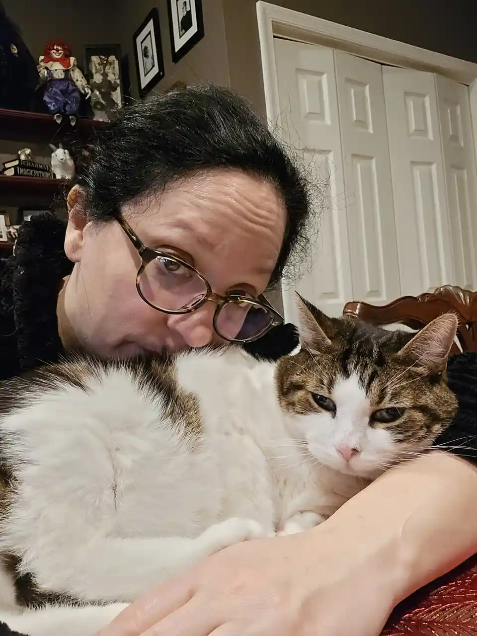 A person with dark hair and glasses leans close to a fluffy white and brown cat they are holding. The cat has a distinctly…
