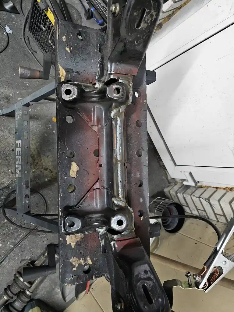 A close-up photo shows a metal car frame section with fresh, shiny welds along its seams, resting on a workbench in a…