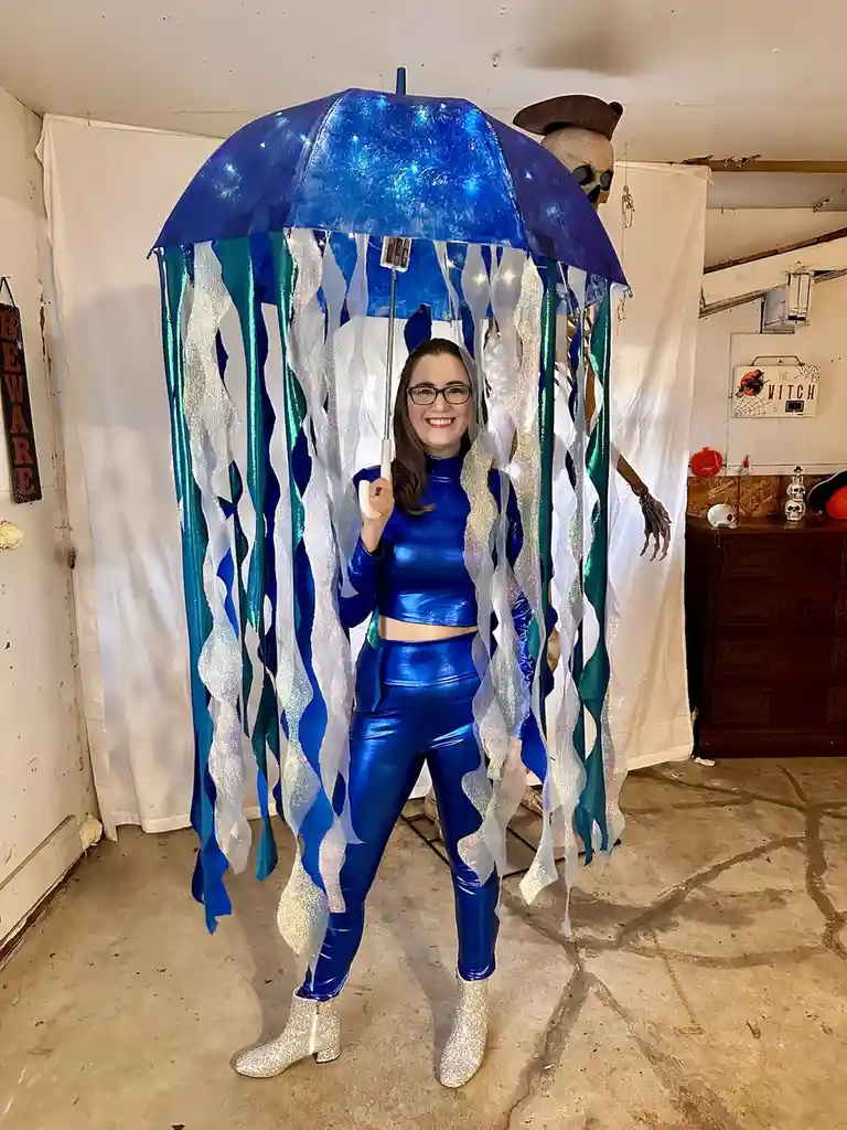 A woman in a shiny blue two-piece outfit and glittery boots stands inside a large blue umbrella decorated with blue, teal…