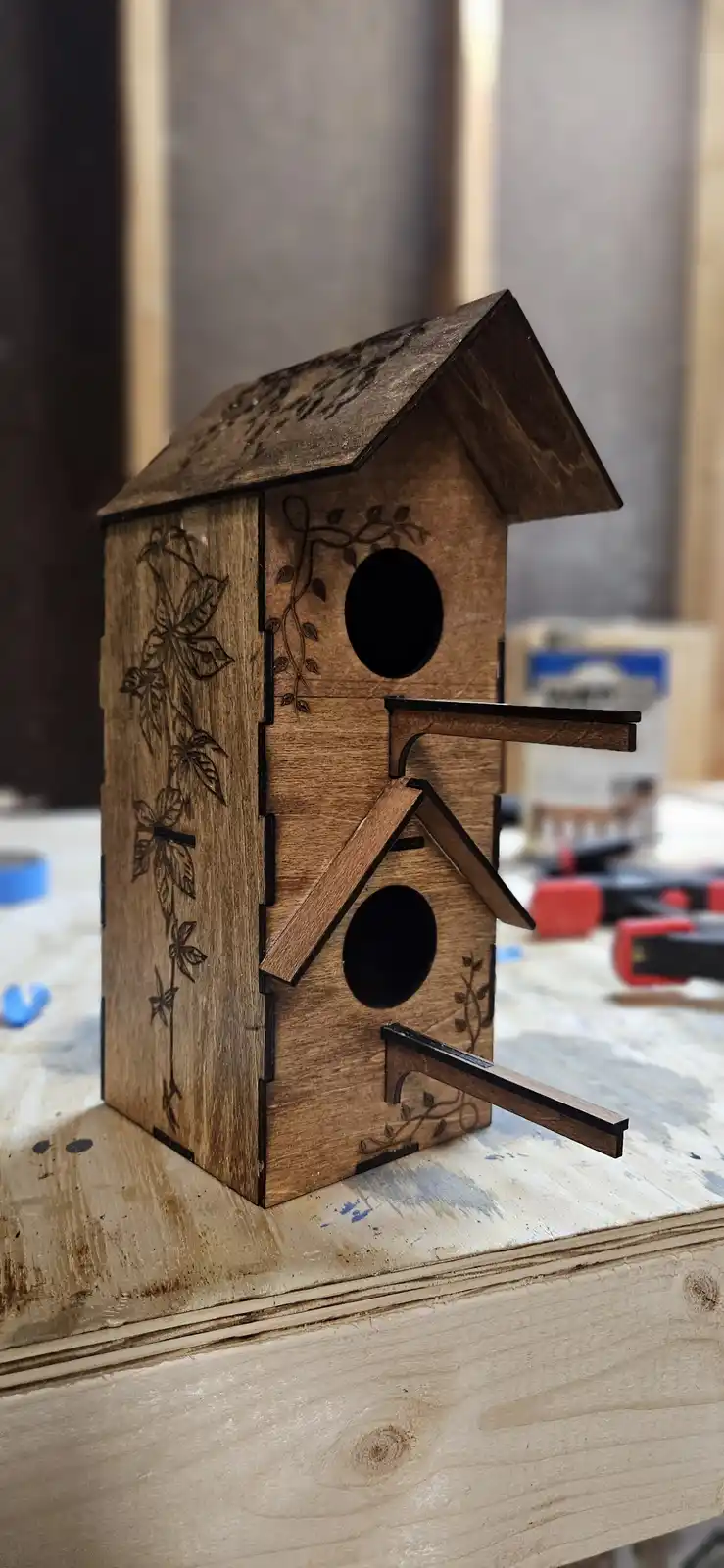 A wooden birdhouse with two circular entry holes and vine etchings sits on a workbench, but its roof and side panels are…