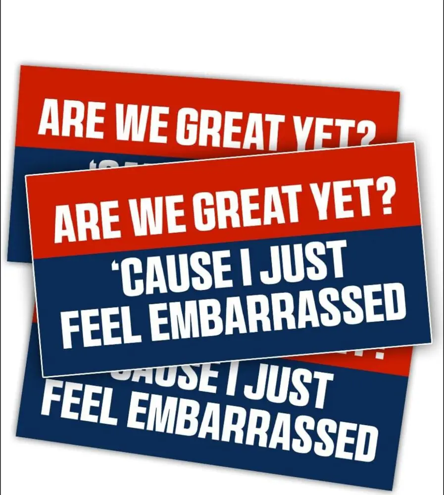 Three identical red-and-blue protest-style signs are stacked, each displaying the text 'ARE WE GREAT YET?' in white bold…