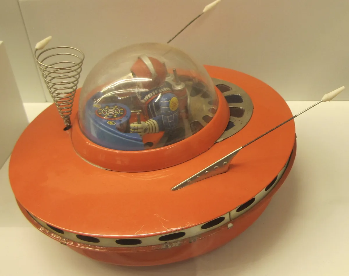A vintage orange toy spaceship with a clear dome containing a small astronaut figure, featuring antennae and a spring-like…