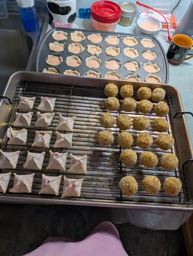 A kitchen counter shows three trays of prepared appetizers: a muffin tin with mini tart shells filled with pink sauce, a…
